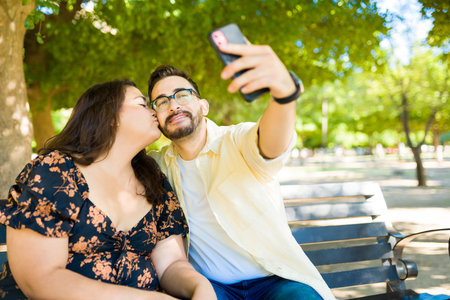 Loving big girlfriend kissing her handsome boyfriend on the cheek while taking a selfie togetherの写真素材