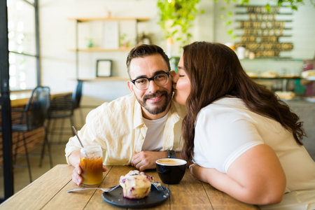 Handsome happy man getting a kiss on the cheek from his plus size girlfriend at the coffee shopの写真素材