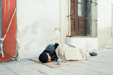 Cold beggar homeless man covered in a blanket sleeping on the street with a please help cardboard signの写真素材