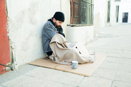 Sad miserable homeless man using a blanket feeling cold in the streets while begging for moneyの写真素材
