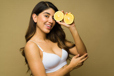 Healthy beautiful mexican woman smiling and taking vitamin C and eating orange fruit for a good skin care routine in front of a studio backgroundの写真素材