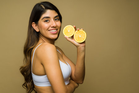 Profile of a cheerful young woman wearing a white bra taking vitamin c and eating orange fruit for good health and clear skinの写真素材