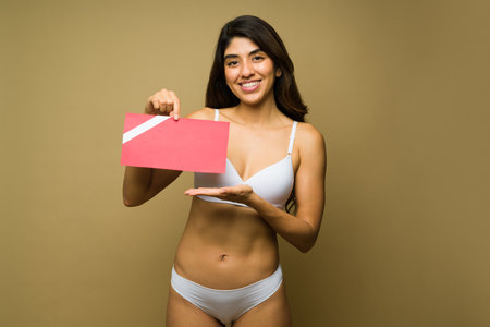 Portrait of a cheerful beautiful woman in underwear holding showing a gift card for hair removal or cosmetic procedureの写真素材