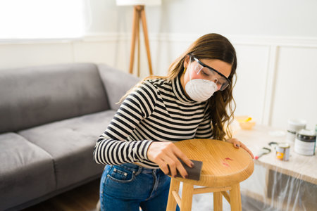Young woman with safety glasses and face mask using sanding paper on a stool furnitureの写真素材
