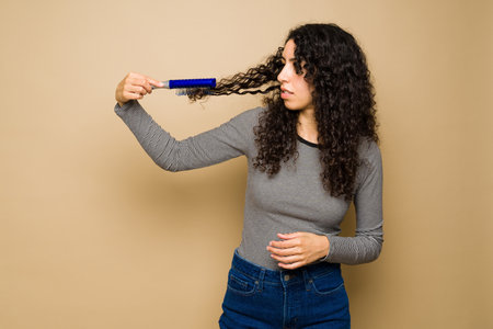 Worried latin young woman with curly hair combing her hair and suffering from tangled hair or frizzの写真素材