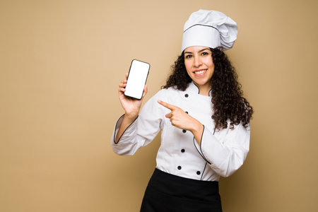 Attractive woman chef or cook smiling while pointing to her smartphone for a food delivery app or social mediaの写真素材