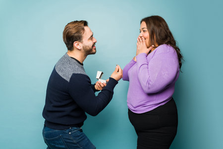 Romantic caucasian man in love proposing with a diamond ring to her big young girlfriend looking surprisedの写真素材