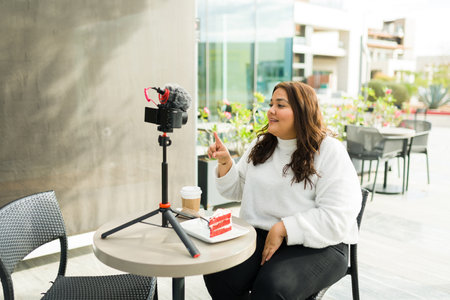 Latin big woman working as video blogger or influencer and filming social media content with a camera trying a cake and coffee at a cafeの写真素材
