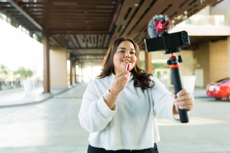 Beautiful hispanic big woman smiling while putting on lipstick and making a video review about makeup productsの写真素材