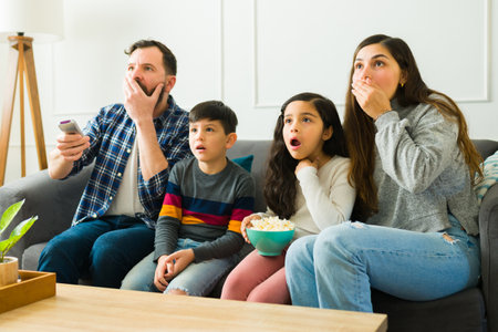 Surprised parents and kids watching a horror movie together and having fun eating popcorn