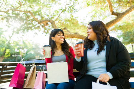 Attractive hispanic friends drinking coffee and smiling at the park after going shopping