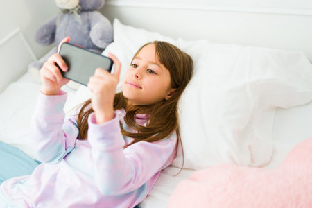 Beautiful caucasian child waking up in the morning playing videogames on the smartphone while in bed