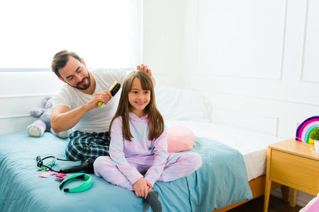 Loving dad brushing the hair of her happy little girl sitting in bed in pajamas while getting readyの写真素材