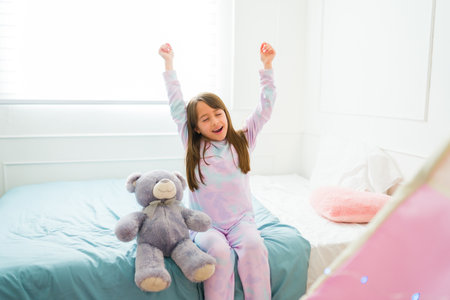 Cute little girl stretching and yawning waking up in the morning with her teddy bear in the bedroomの写真素材