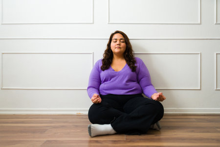 Relaxed obese woman meditating and doing breathing exercises while sitting on the floor at home...