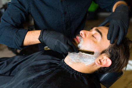 Close up of a barber using a razor blade to shave the beard of a relaxed caucasian man at the salonの写真素材