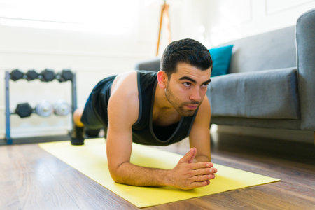 Determined latin man doing plank exercises and working out with motivation in his living room home