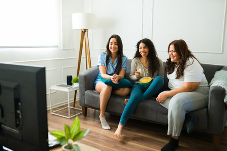 Happy female friends laughing and eating snacks while relaxing together watching a movie in the living room during a sleepoverの写真素材