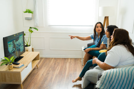 Latin female friends relaxing on the couch looking surprised while watching movies together in the...