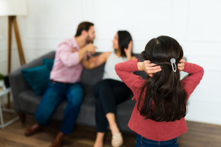 Rear view of an upset little girl and daughter covering her ears while looking at her angry parents fighting at homeの写真素材