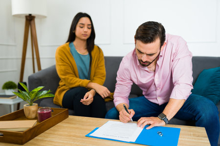 Caucasian angry man signing the divorce papers with a sad hispanic woman sitting on the sofa at homeの写真素材