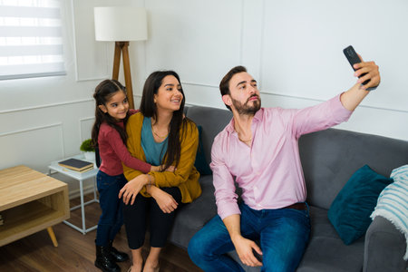 Caucasian father taking a selfie with his beautiful happy family while sitting together on the sofa in the living roomの写真素材