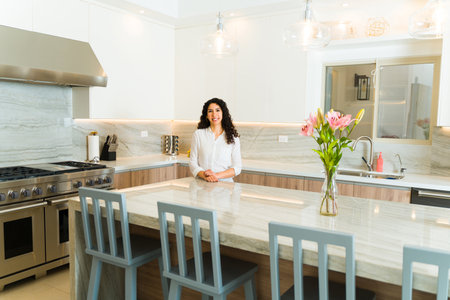 Happy latin young woman making eye contact smiling standing in her beautiful luxury kitchen ready to start cookingの写真素材