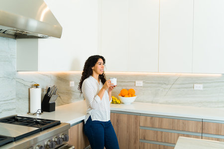 Smiling happy woman standing drinking a cup of coffee in the morning in her beautiful sunny luxury kitchenの写真素材