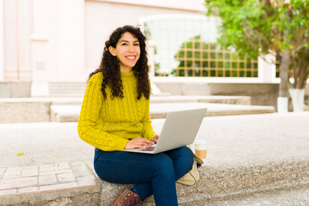 Beautiful happy woman with curly hair smiling and working on her laptop outdoors doing remote workの写真素材