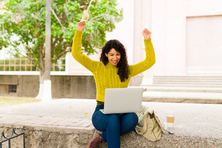 Happy beautiful woman celebrating looking excited while working outdoors on her laptop and receiving good newsの写真素材