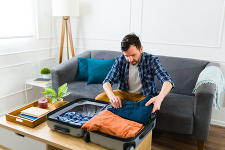 Organized caucasian man at home packing clothes in his suitcase and preparing to go on a travel or vacationsの写真素材