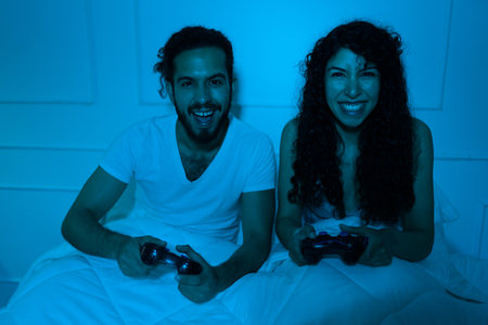 Cheerful beautiful young couple laughing and having fun while playing videogames in bed before...