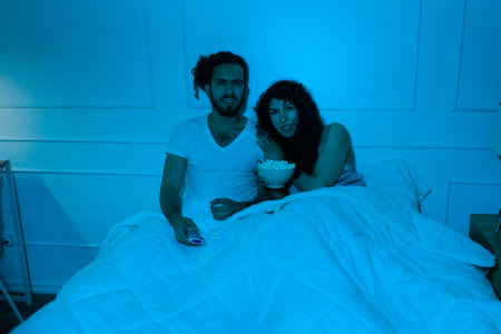 Scared hispanic couple watching a horror movie at night and feeling afraid in bed while eating popcornの写真素材