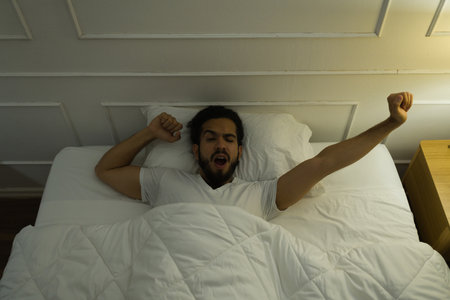 High angle of a relaxed latin man stretching his after waking up in bed under the coversの写真素材