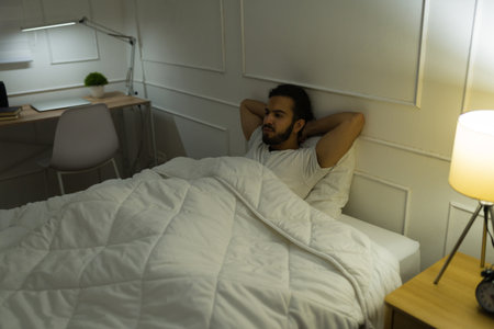 Attractive hispanic man sitting up in bed and suffering from sleeping problems and insomnia late at night in his bedroomの写真素材