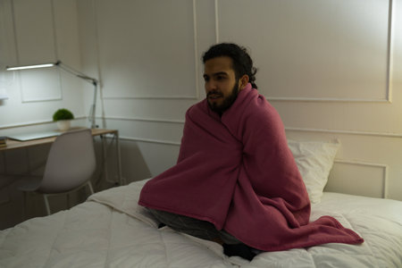 Upset young man wrapped in a blanket and covering from the cold in bed during the winter having sleeping problemsの写真素材