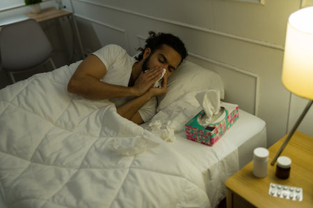 Sick latin man under the covers resting in bed and blowing his nose while suffering from a bad coldの写真素材
