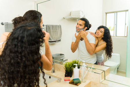 Cheerful young man shaving his beard and talking with his girlfriend while getting ready in the morningの写真素材