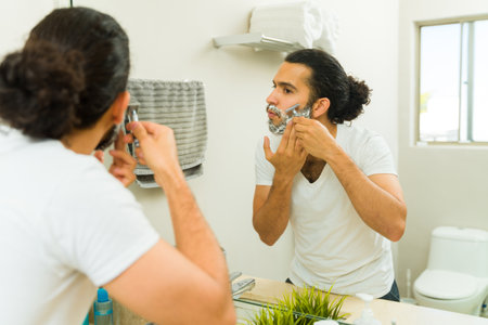 Handsome man in the bathroom shaving his beard with a razor and using grooming productsの写真素材
