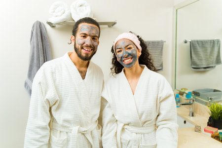 Young woman and man smiling in white robes using face mud mask doing their skin care routine during a relaxing spa day at homeの写真素材