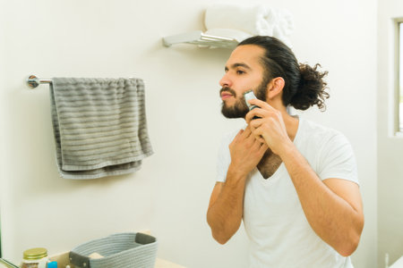 Attractive young man in his 20s using a trimmer and grooming his beard in the bathroom mirrorの写真素材