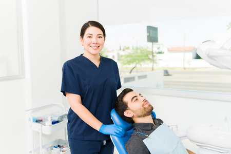 Beautiful caucasian female dentist smiling in scrubs and making eye contact while doing a patient examinationの写真素材