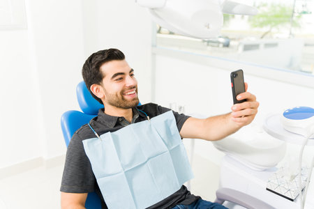 Latin man smiling looking happy taking a selfie with his smartphone to post on social media while at the dentist for an examinationの写真素材