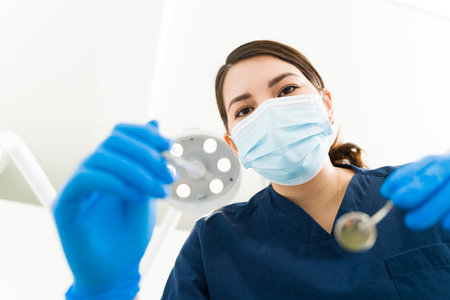 Professional female caucasian dentist with a face mask wearing scrubs and gloves making eye contact while doing a dental examination of a patientの写真素材