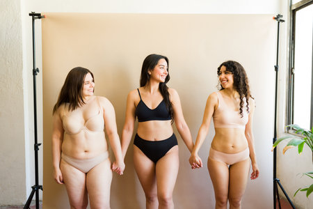 Loving young women with diverse bodies in underwear holding hands and laughing together looking feminine and with a lot of self-loveの写真素材