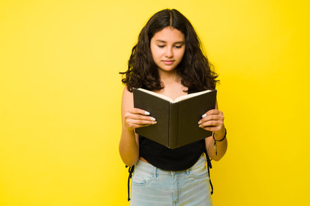 Smart teenage girl reading an interesting book and looking relaxed in a copy space yellow ad in a...