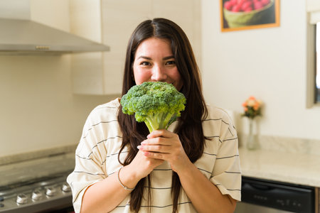 Attractive vegan woman making eye contact and smiling while showing a green vegetable broccoli ready to cook healthy foodの写真素材