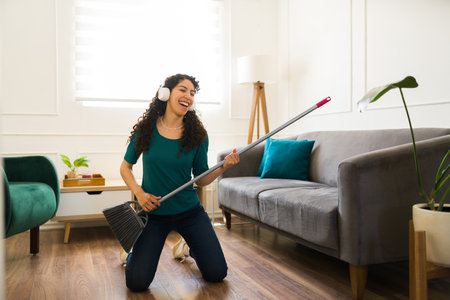 Excited young woman doing housework and cleaning chores while having fun and sweeping the living room listening to musicの写真素材