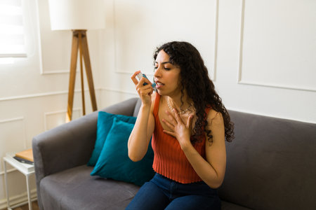 Sick hispanic young woman using an inhaler while having an asthma attack and breathing problems in...