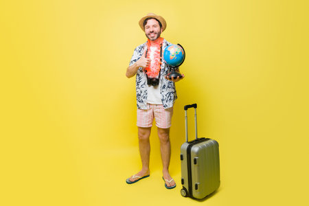 Full length of a mid-adult tourist man smiling holding a globe while ready to travel for summer vacations against a yellow backgroundの写真素材